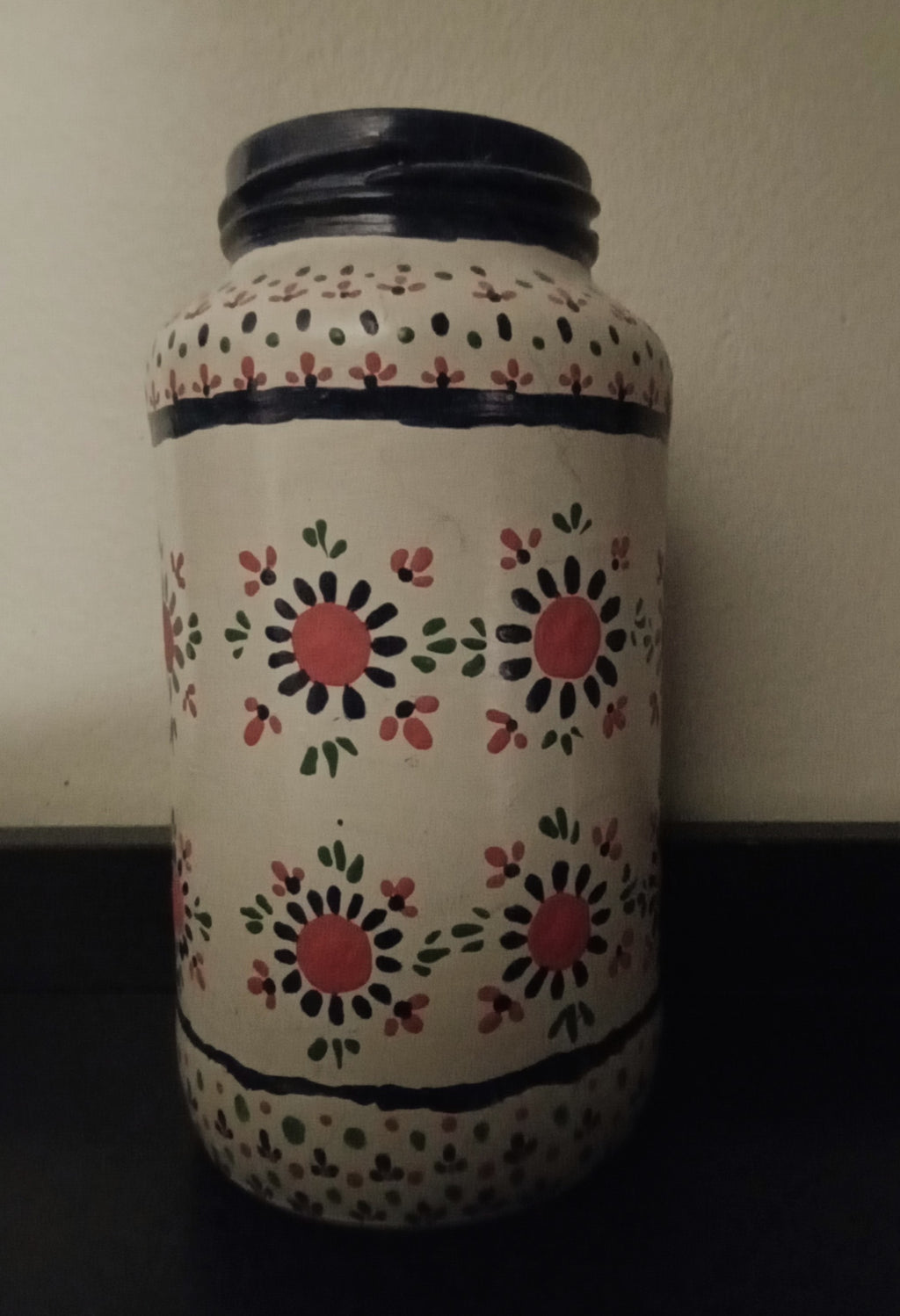 A hand-painted glass jar featuring a cream-colored body decorated with red, black, and green floral motifs and blue horizontal stripes.