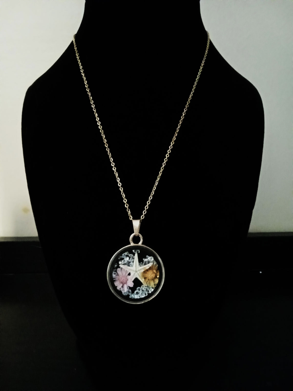 A gold-toned chain necklace with a round pendant. The pendant features a   starfish and small dried flowers.