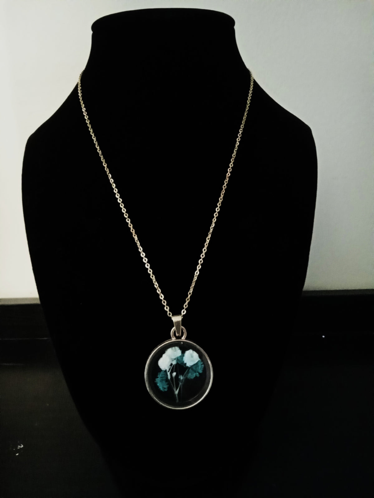 A gold-toned chain necklace with a round pendant displaying a dried floral design in shades of green and white.