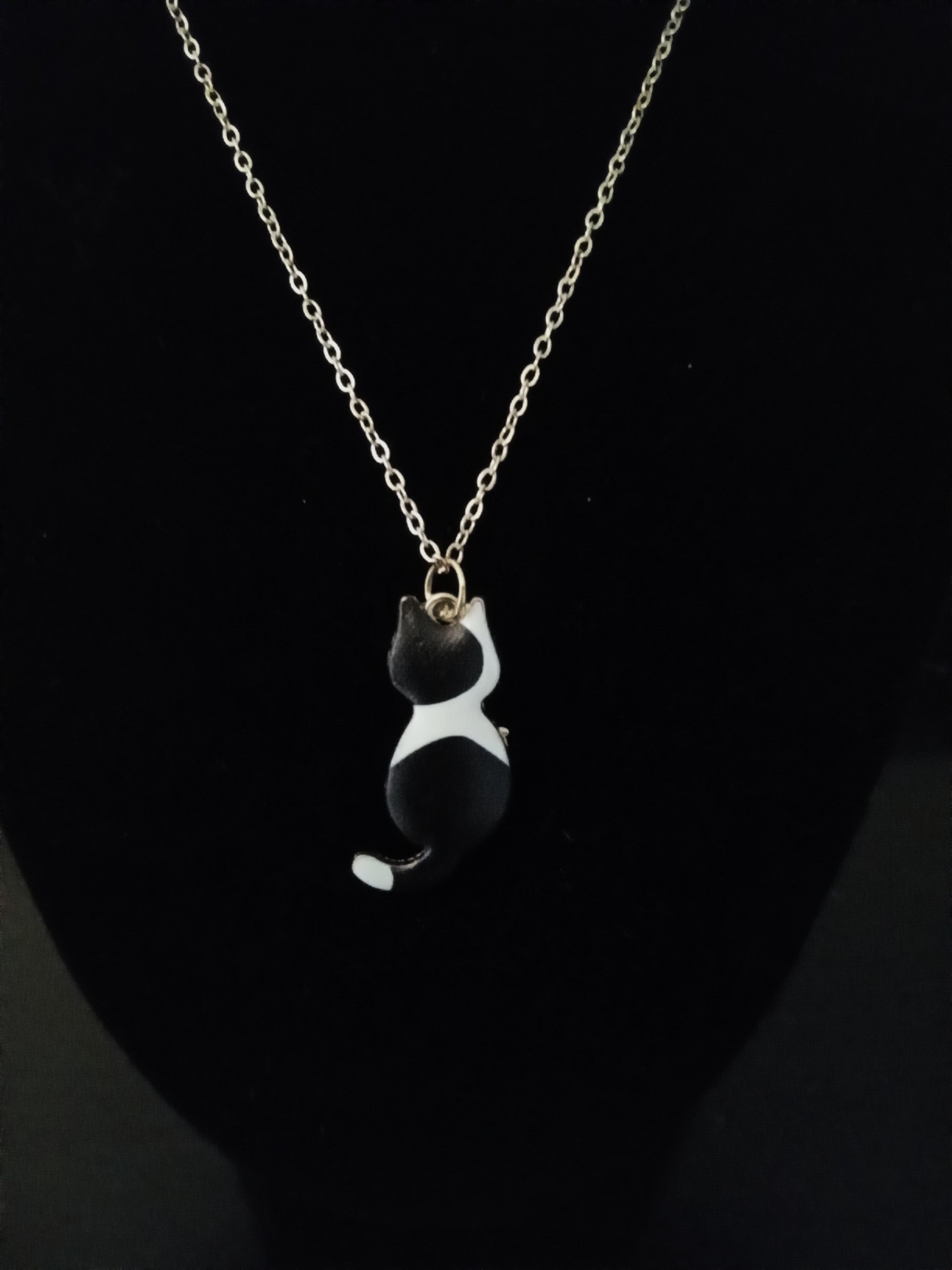A gold chain necklace featuring a small black and white cat pendant, displayed on a black bust.