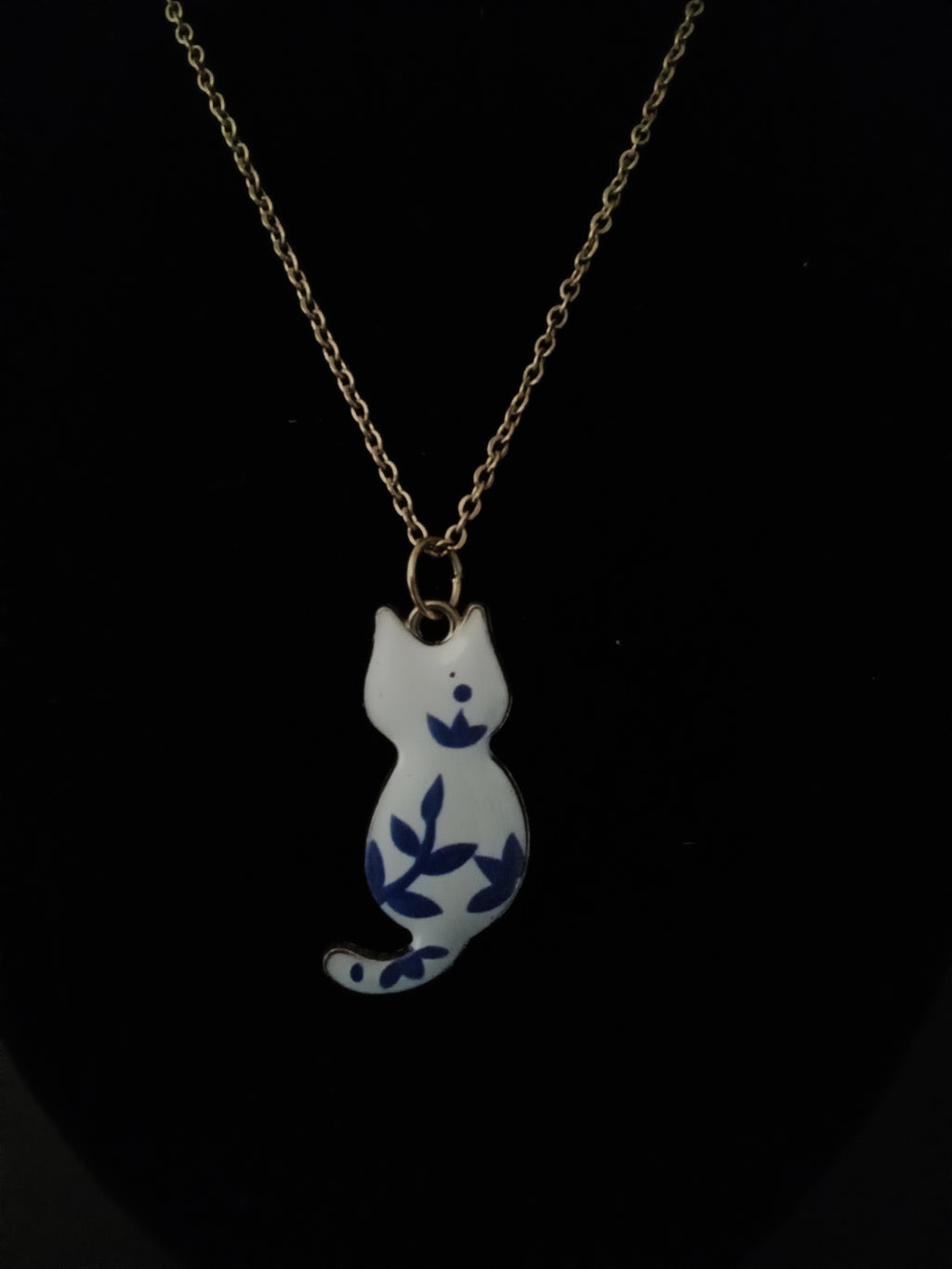 A gold chain necklace featuring a white cat-shaped pendant decorated with blue floral and leaf patterns, displayed against a solid black background.