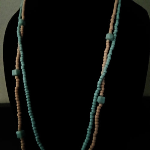 A long beaded necklace displayed on a black mannequin bust. The necklace features two strands of small round beads in light blue and pale peach, separated by larger matching light blue square beads.