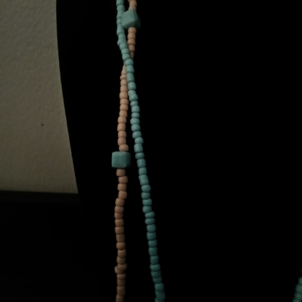 A close-up of a beaded necklace or strand with alternating strands of small round beads in light blue and pale peach, accented by larger light blue square beads, shown against a dark background.