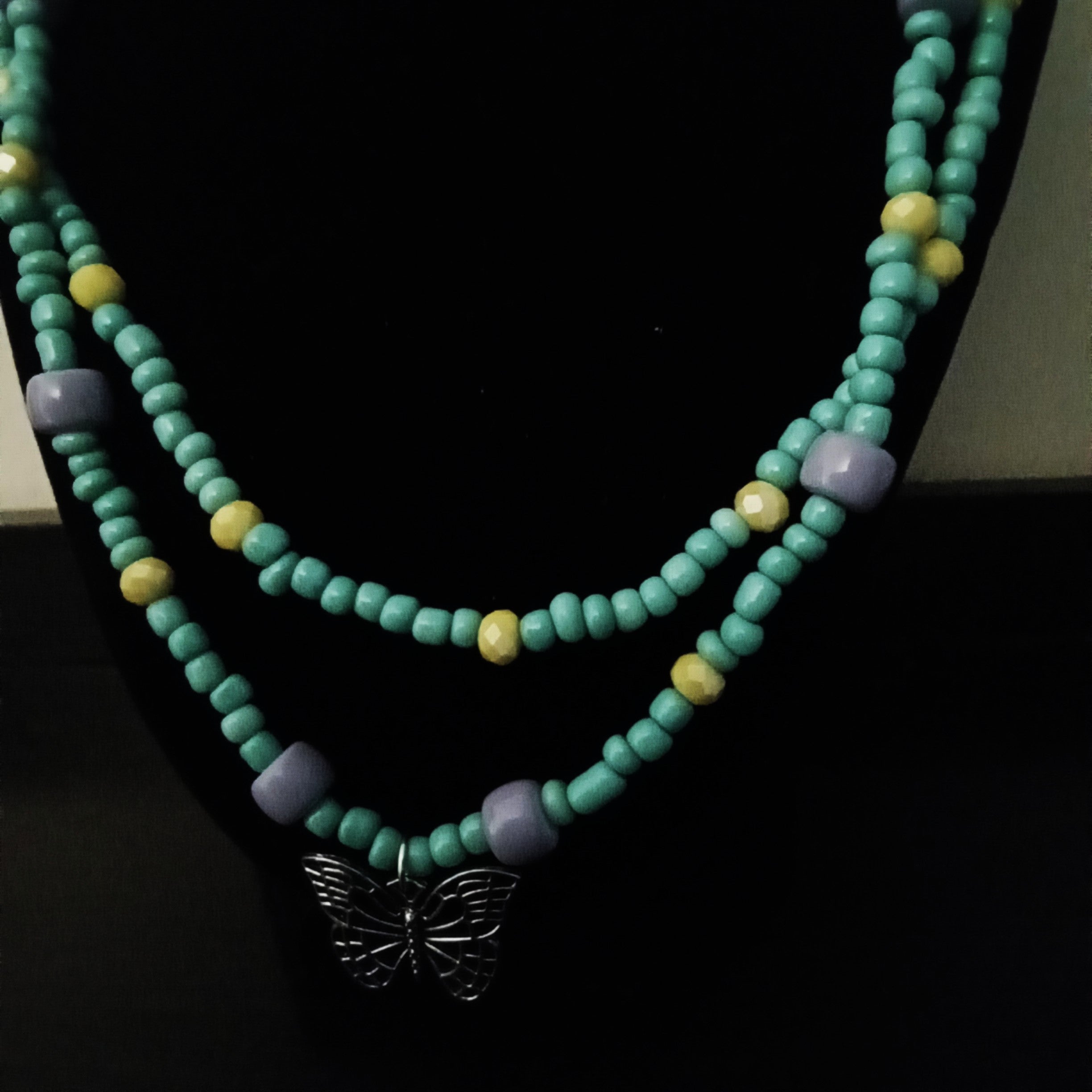 A double-strand beaded necklace displayed on a black jewelry bust. The necklace features turquoise-colored beads, small yellow accent beads, lavender-purple spacer beads, and a silver butterfly pendant at the center.
