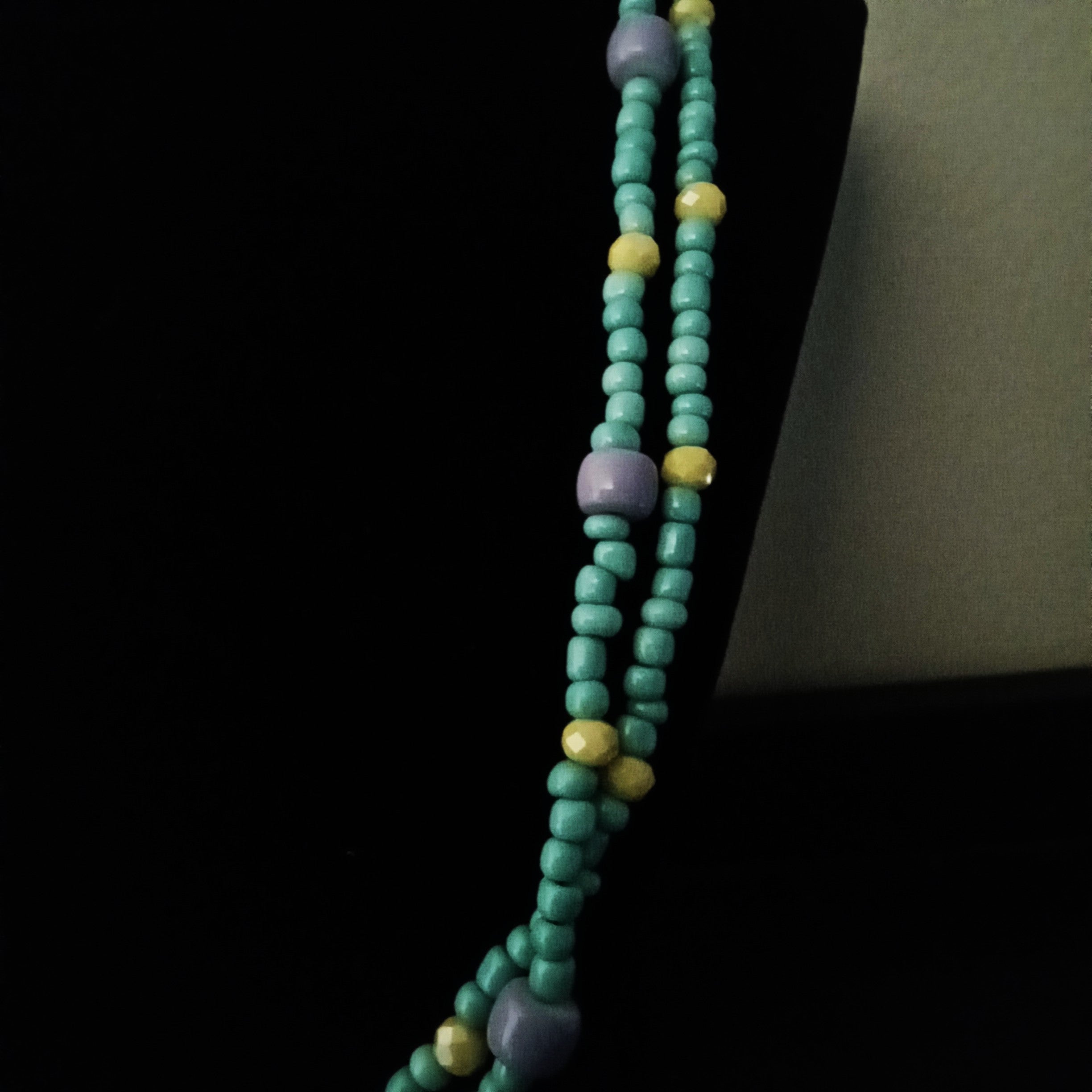 A beaded necklace with turquoise-colored round beads, lavender cylindrical spacer beads, and small yellow round beads, displayed on a black jewelry stand.