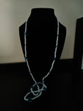 A long, beaded necklace displayed on a black mannequin bust against a dark background. The necklace features light blue seed beads with small blue translucent square beads spaced along the chain.