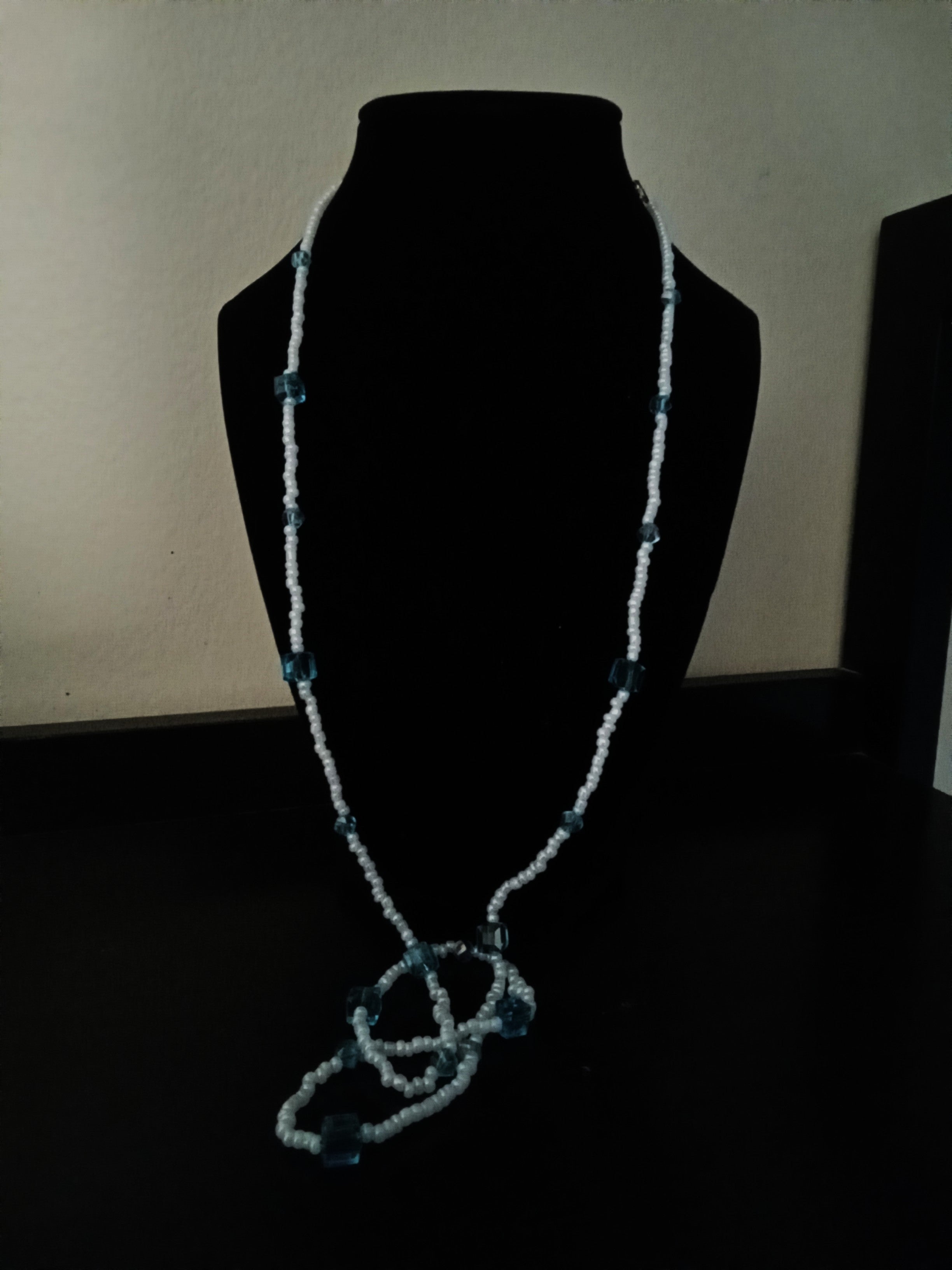 A long, beaded necklace displayed on a black mannequin bust against a dark background. The necklace features light blue seed beads with small blue translucent square beads spaced along the chain.