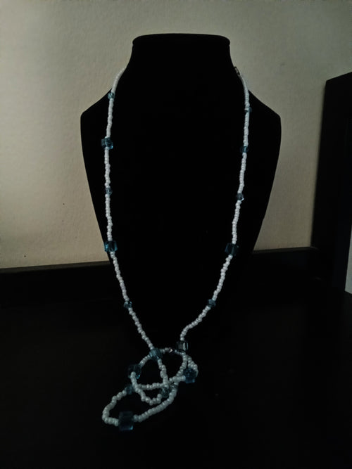 A long, beaded necklace displayed on a black mannequin bust against a dark background. The necklace features light blue seed beads with small blue translucent square beads spaced along the chain.