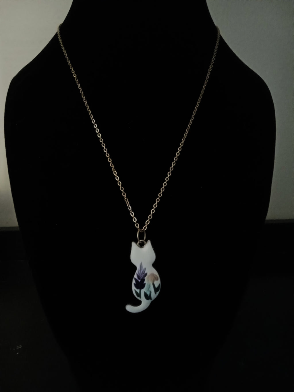 A delicate gold-tone chain necklace with a pendant shaped like a white cat, featuring a purple and green floral design on its body, displayed on a black mannequin bust.