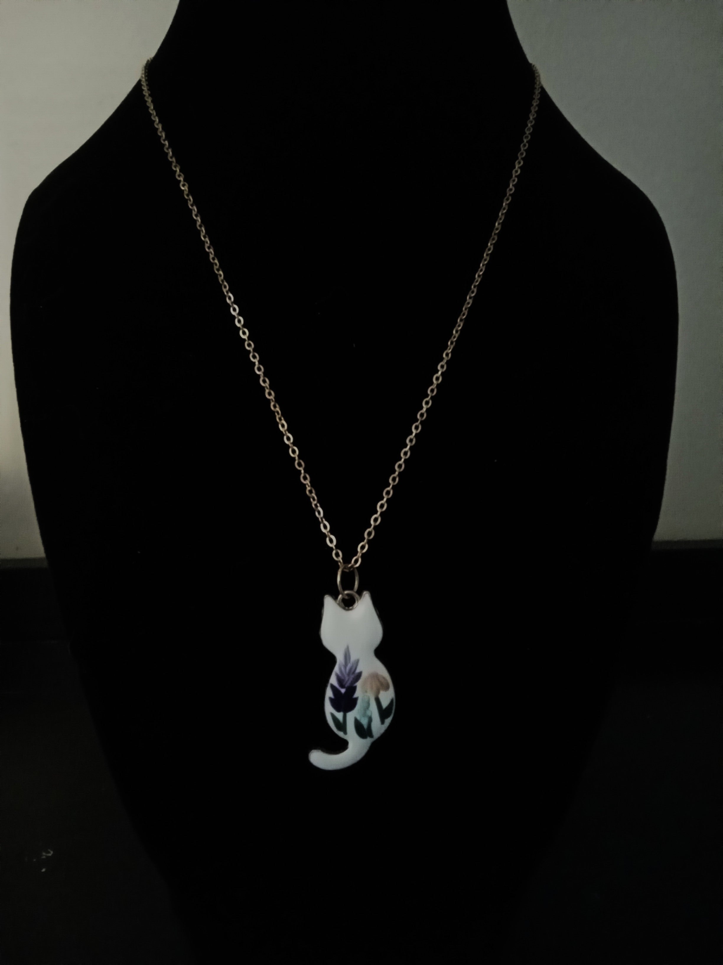 A delicate gold-tone chain necklace with a pendant shaped like a white cat, featuring a purple and green floral design on its body, displayed on a black mannequin bust.