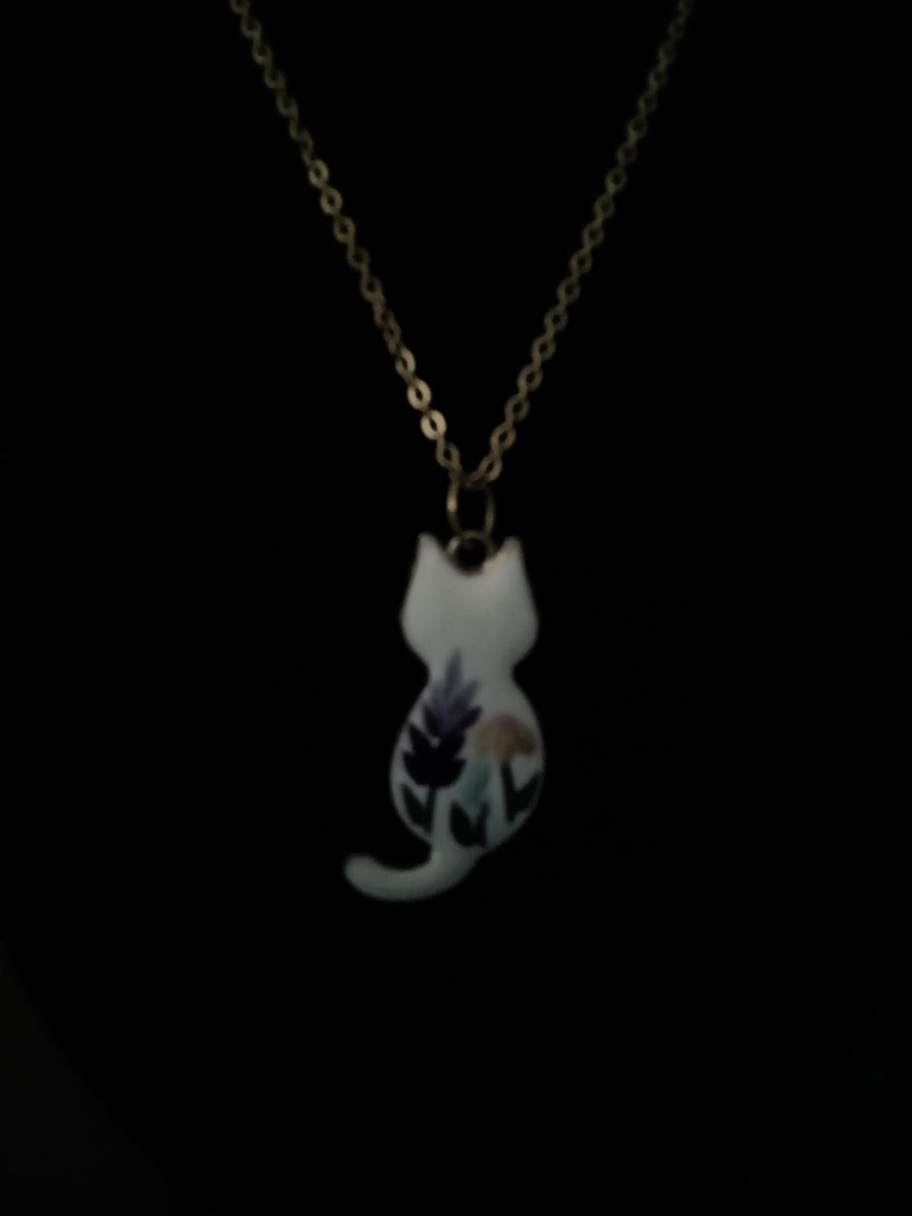 A delicate gold chain necklace featuring a small white cat-shaped pendant decorated with purple and green floral details, shown against a solid black background.