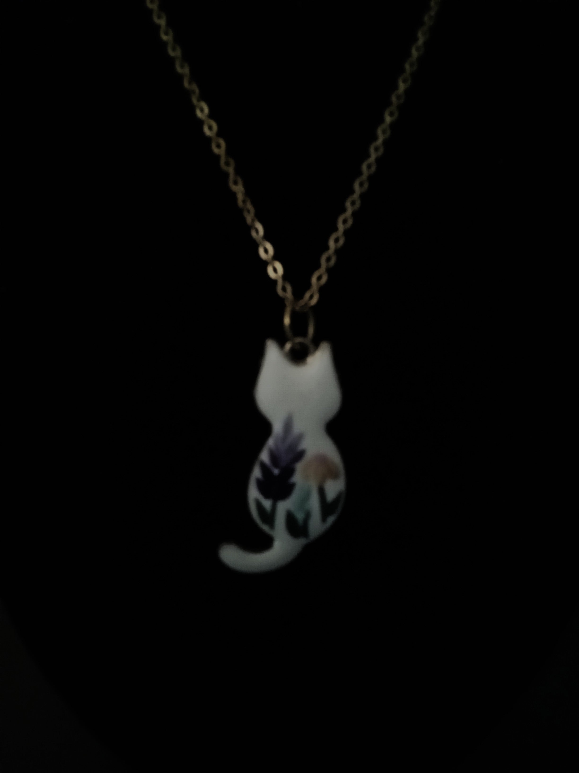 A delicate gold chain necklace featuring a small white cat-shaped pendant decorated with purple and green floral details, shown against a solid black background.