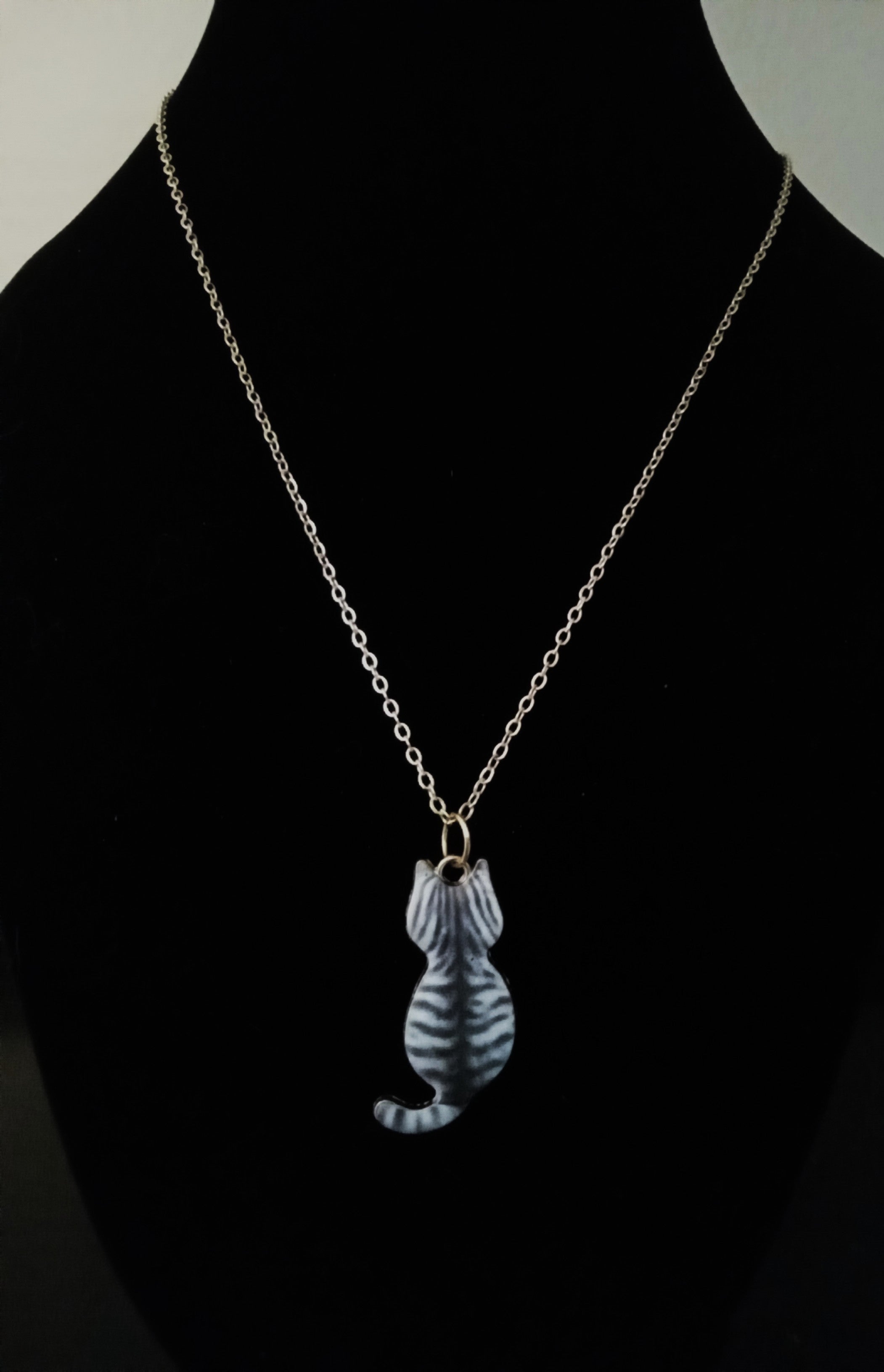 A gold chain necklace with a small cat-shaped pendant featuring gray and white stripes, displayed on a black mannequin bust.