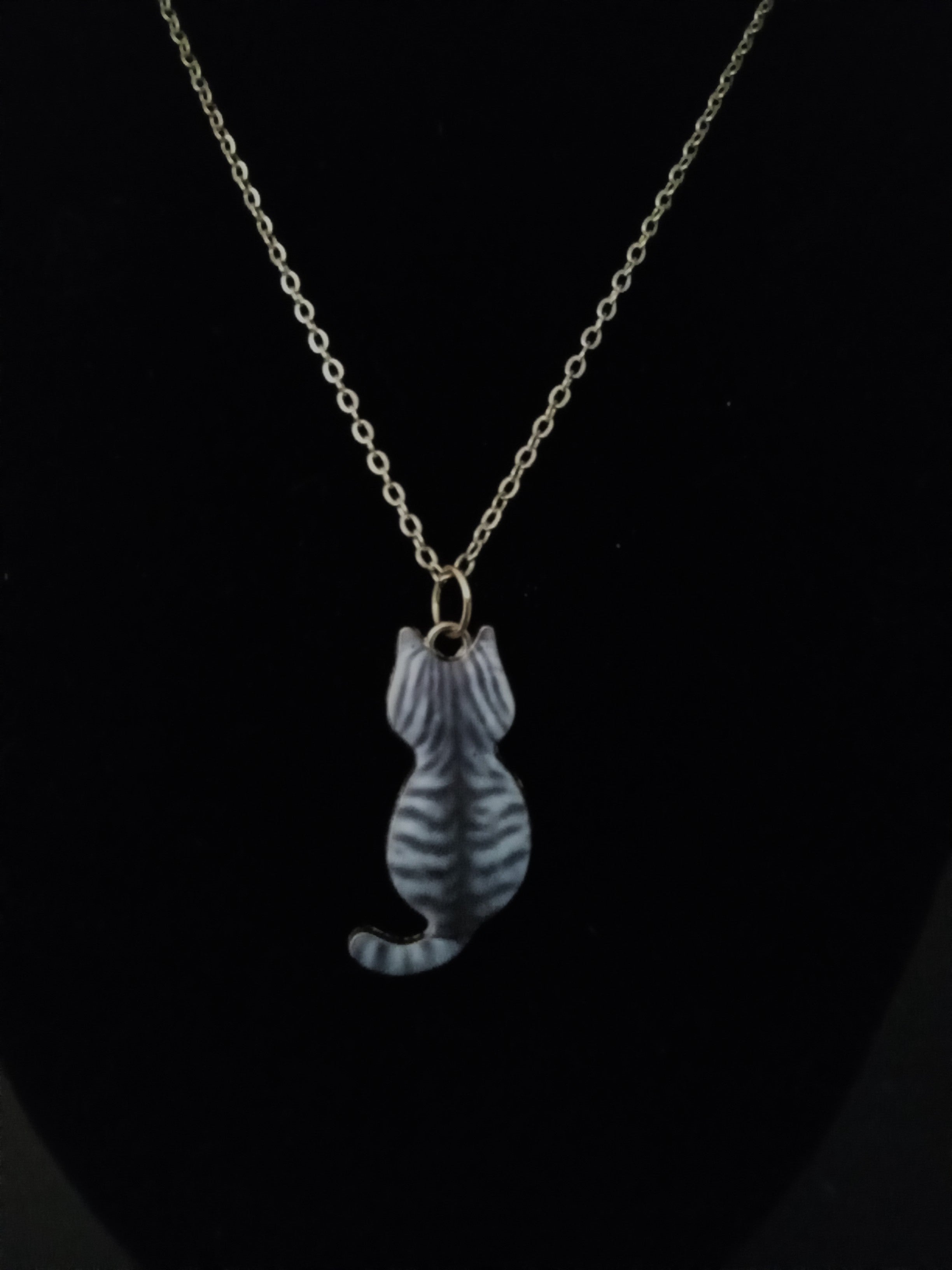 A gold-toned chain necklace with a small cat-shaped pendant featuring gray and white stripes, displayed against a solid black background.
