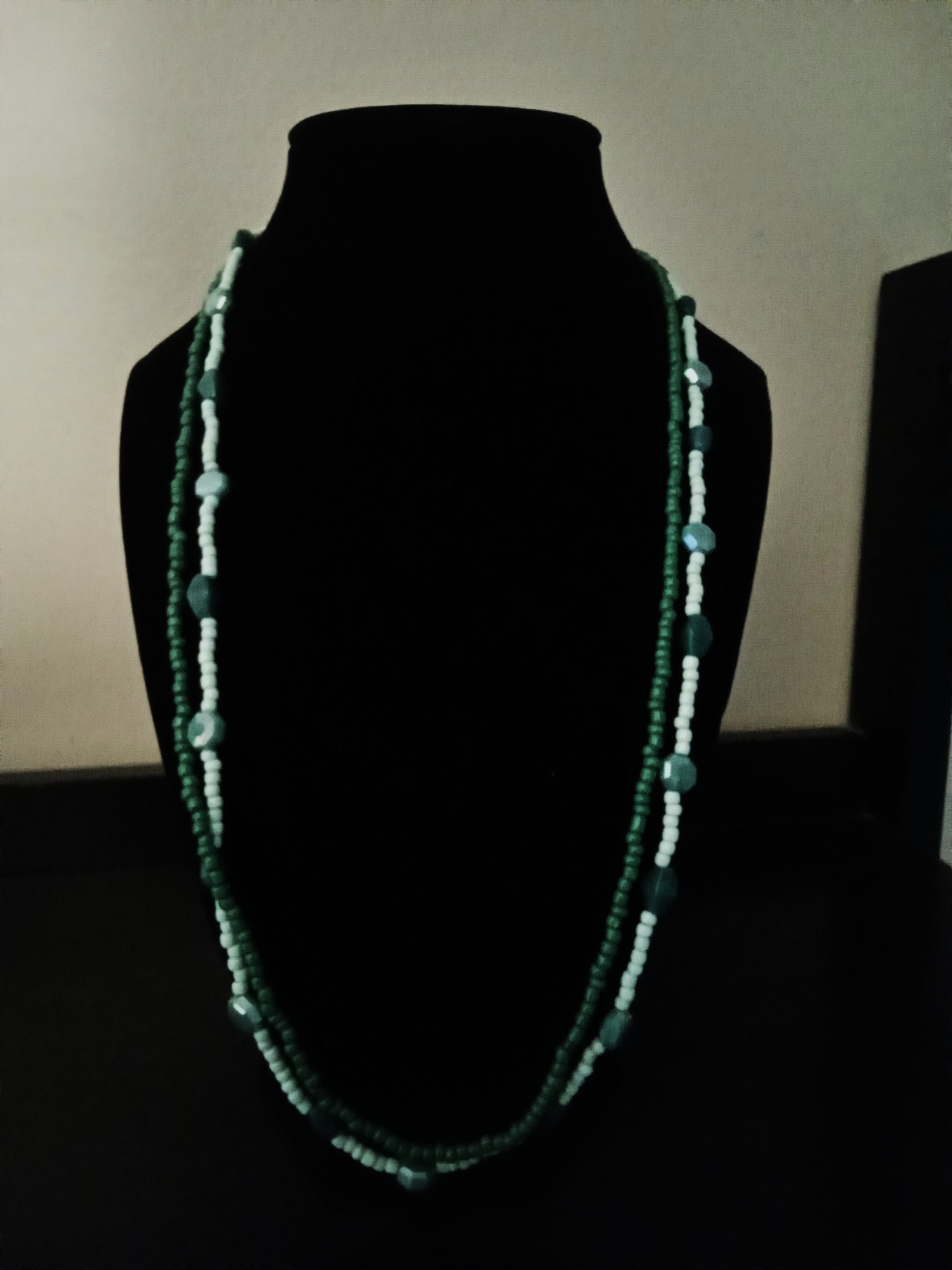 A beaded necklace displayed on a black velvet jewelry bust. The necklace features dark green and light green beads arranged in two parallel strands, with small dark green spacer beads throughout.