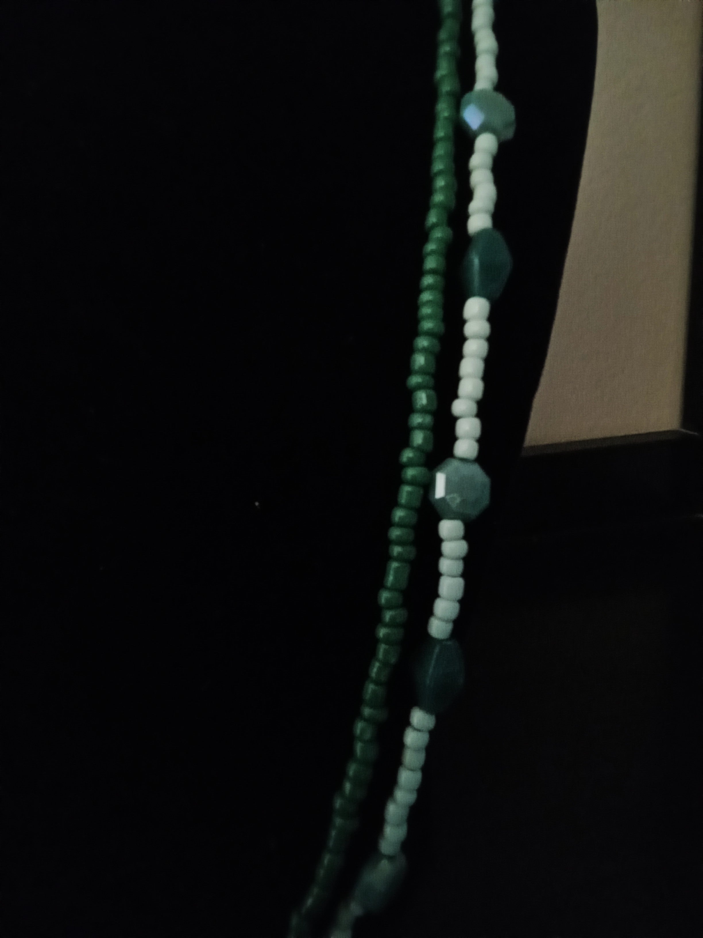A close-up of a beaded necklace draped vertically against a black background, featuring dark green faceted beads and light green round beads.