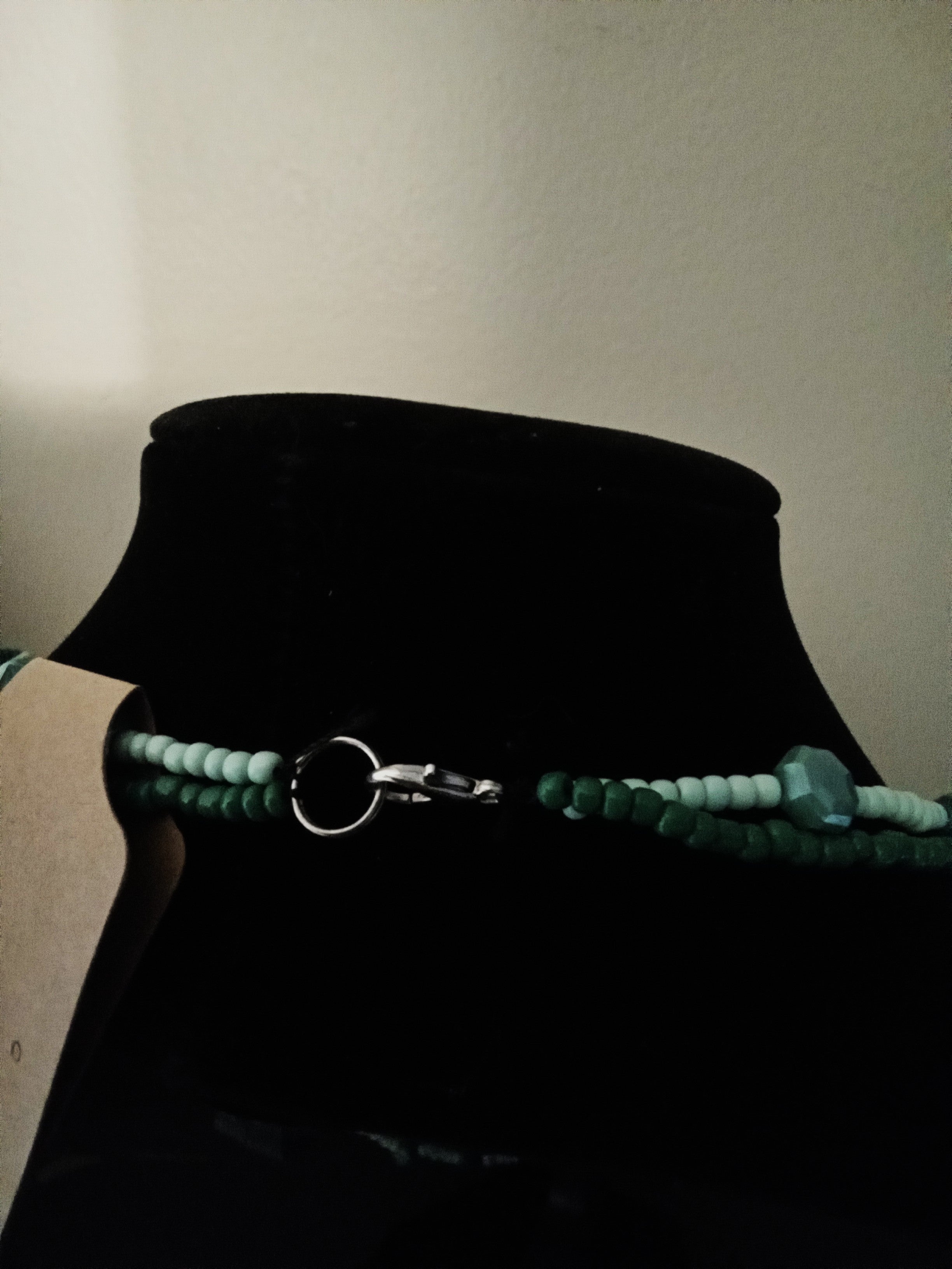 A beaded necklace with dark green and light mint green beads, featuring a clasp, displayed on a black velvet jewelry bust.