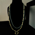 A long, beaded necklace displayed on a black mannequin bust against a dark background. The necklace features multiple strands of small beads in sage green, yellow, and dark green.