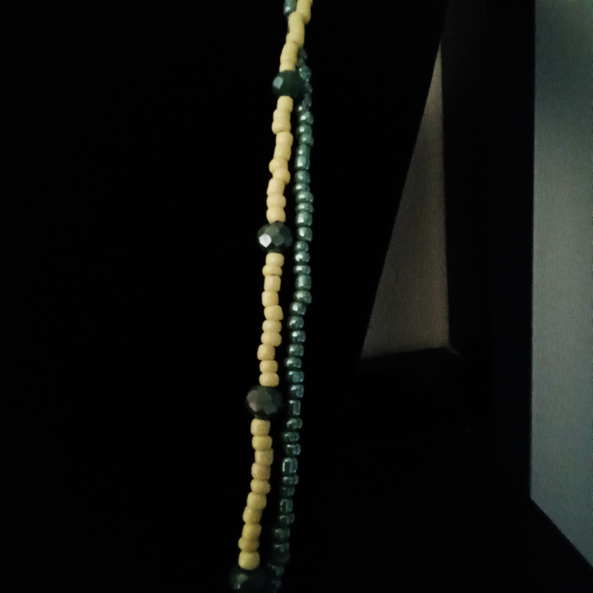 A close-up of a beaded necklace featuring alternating vertical columns of yellow beads and dark green faceted beads, set against a very dark background.