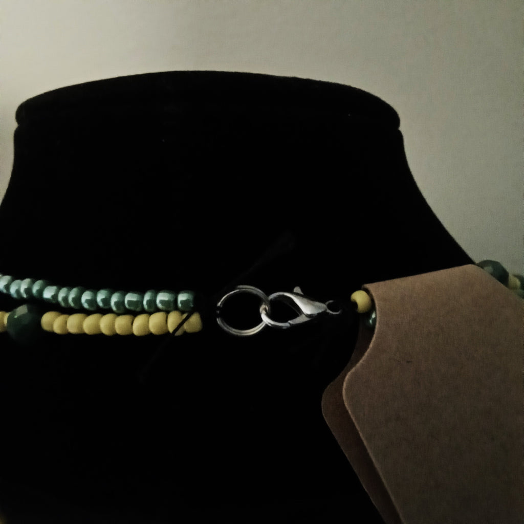 A close-up of a beaded necklace displayed on a black velvet jewelry bust. The necklace features two strands: one with iridescent green beads and one with yellow beads, joined by a silver lobster clasp.