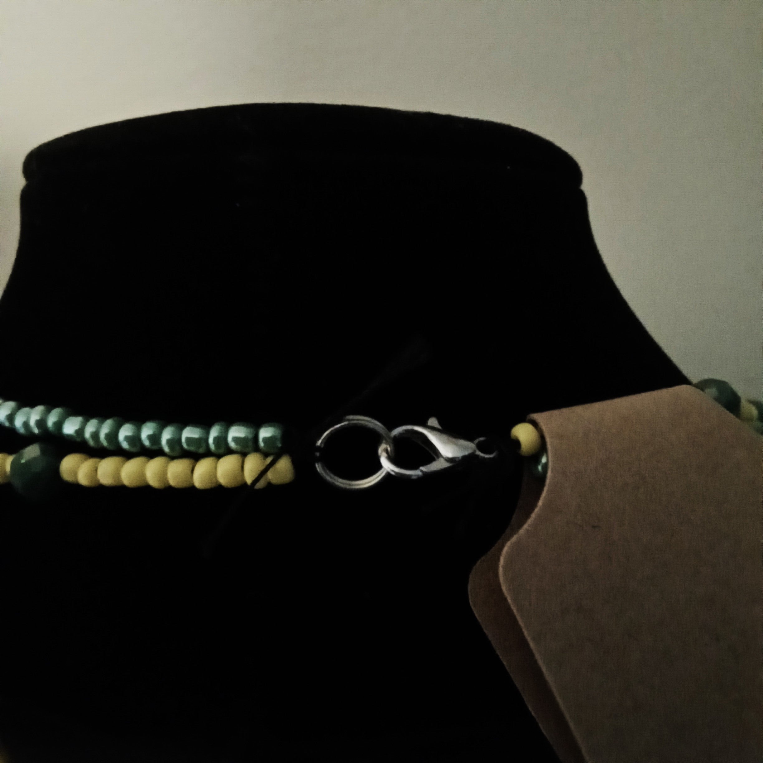 A close-up of a beaded necklace displayed on a black velvet jewelry bust. The necklace features two strands: one with iridescent green beads and one with yellow beads, joined by a silver lobster clasp.