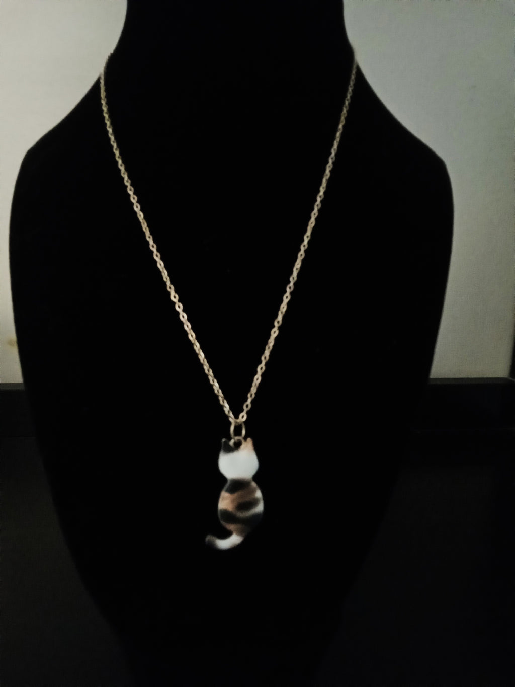 A gold chain necklace with a cat-shaped pendant featuring a white, black, and brown marbled pattern, displayed on a black jewelry bust.