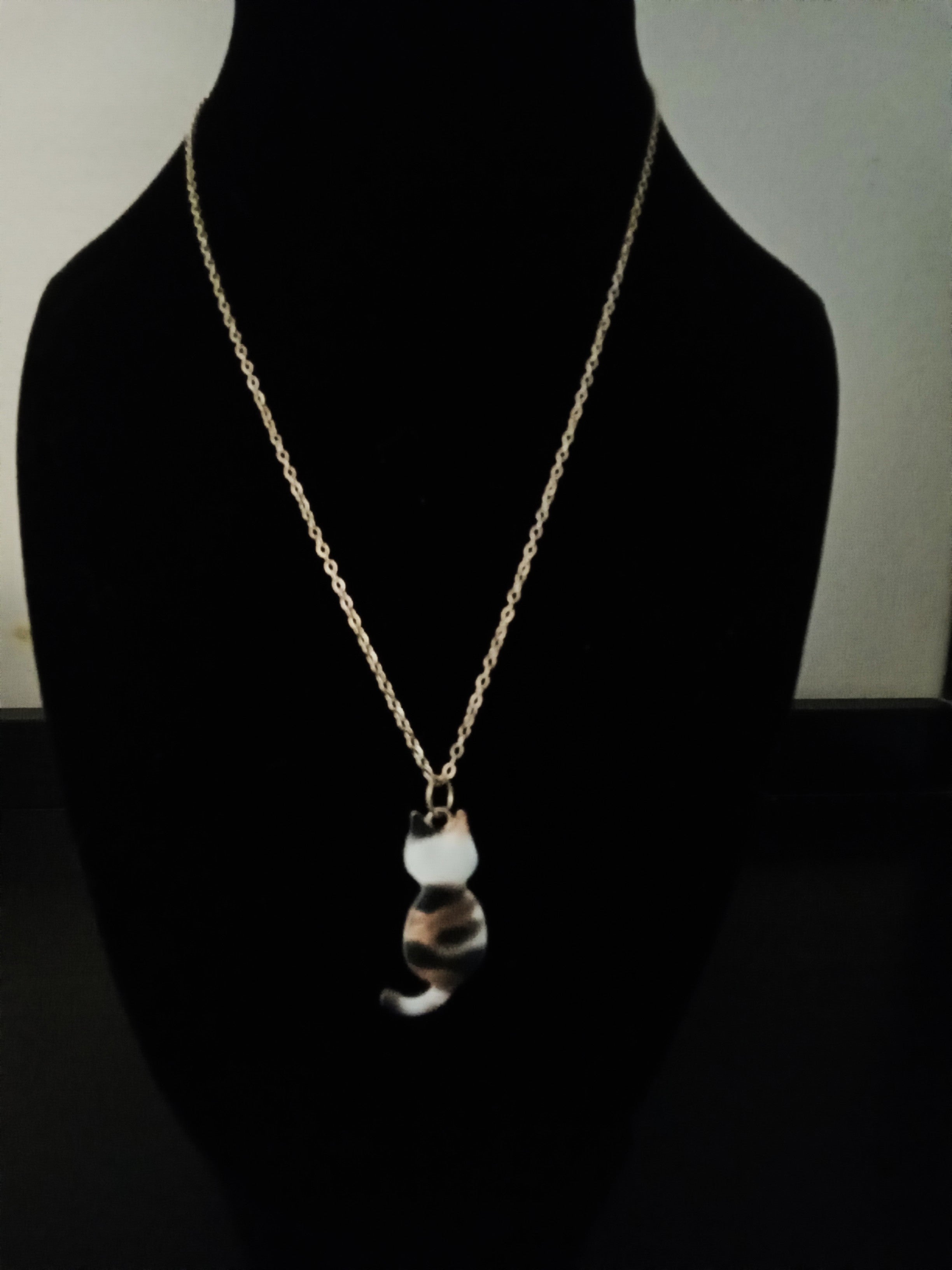 A gold chain necklace with a cat-shaped pendant featuring a white, black, and brown marbled pattern, displayed on a black jewelry bust.