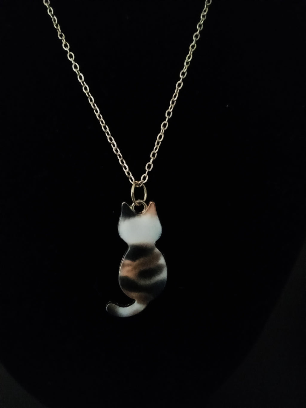 A gold-tone chain necklace with a small cat-shaped pendant featuring a marbled pattern of black, white, and brown, displayed against a solid black background.