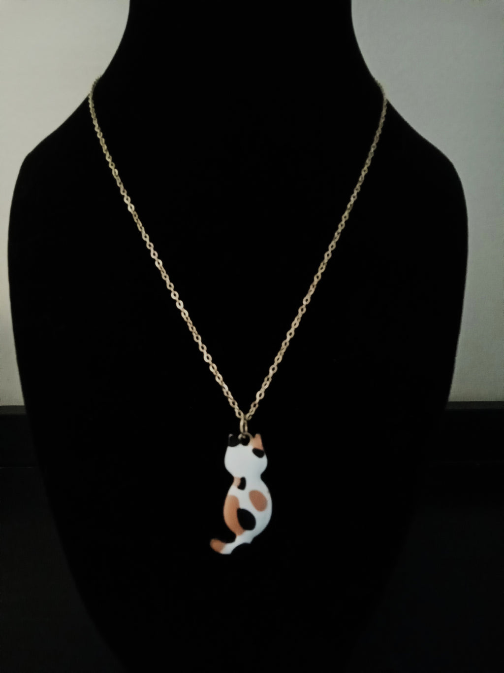 A gold chain necklace with a small cat-shaped pendant, displayed on a black mannequin bust. The pendant is white with orange and black spots, resembling a calico cat.