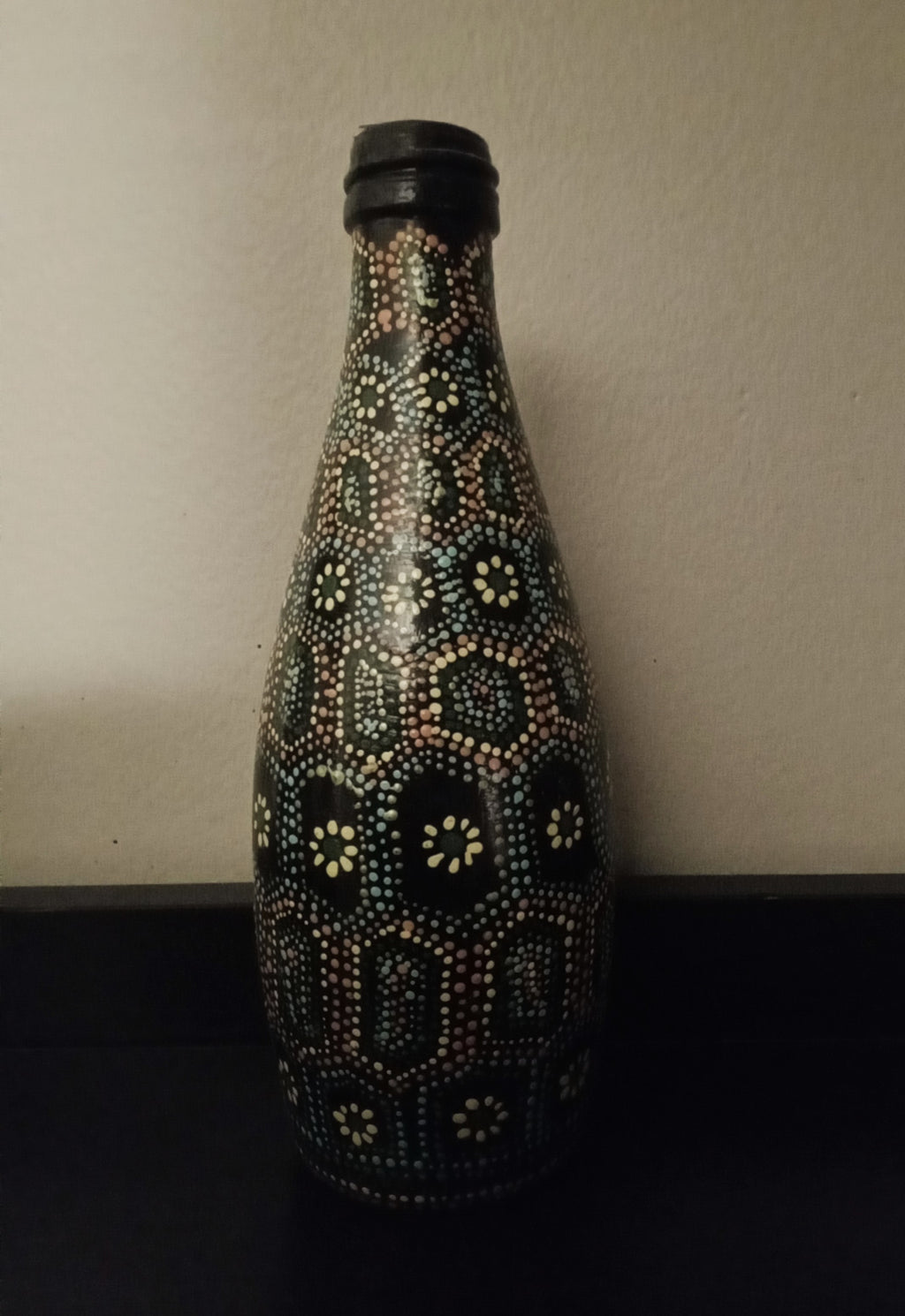 A black glass bottle decorated with intricate hand-painted Aboriginal-style dot art patterns in yellow, green, orange, and light blue.
