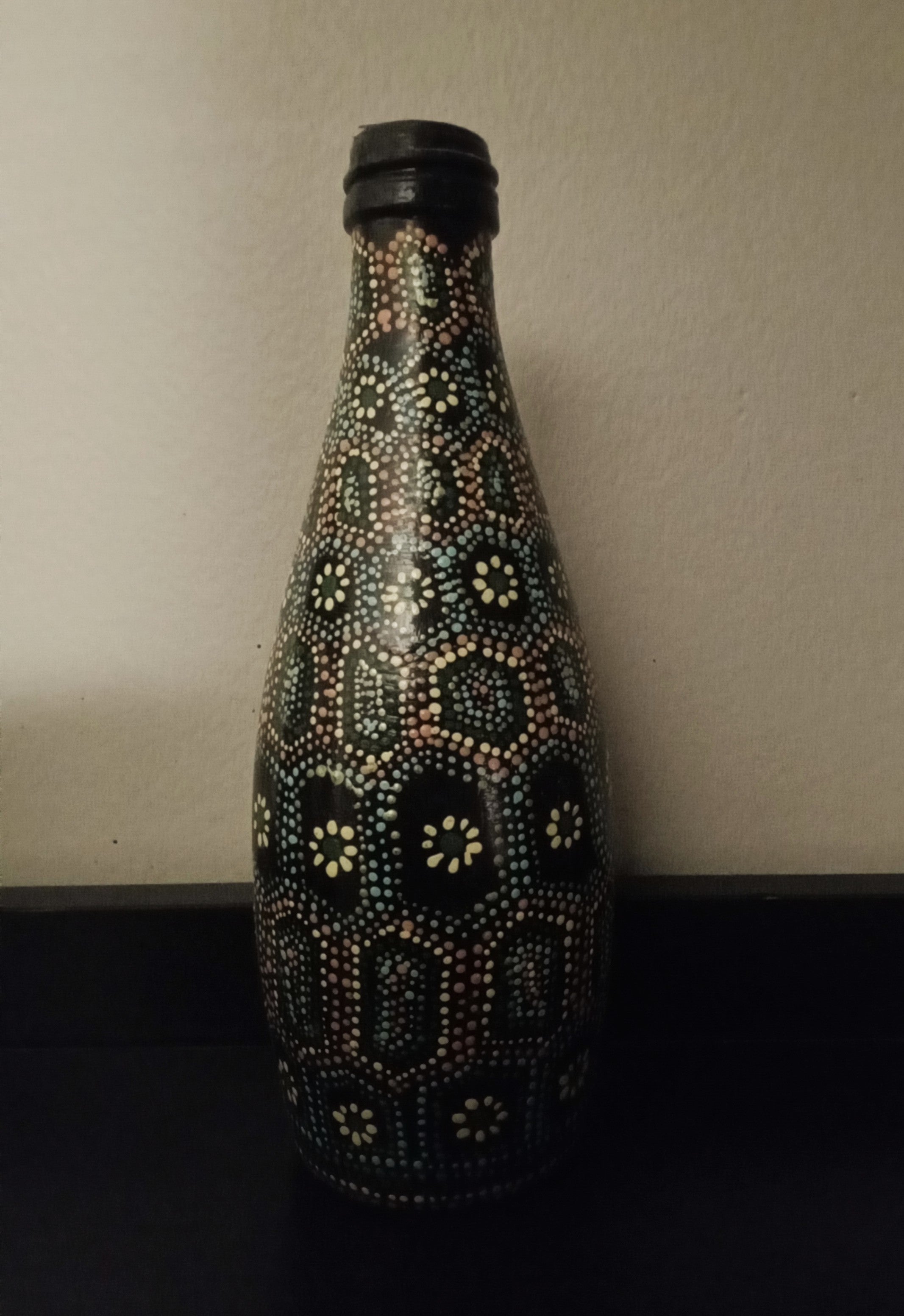 A black glass bottle decorated with intricate hand-painted Aboriginal-style dot art patterns in yellow, green, orange, and light blue.