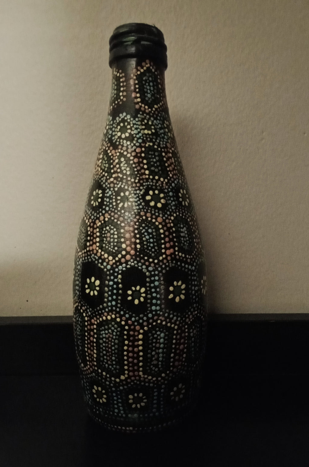 A black glass bottle decorated with intricate hand-painted Aboriginal-style dot art patterns in yellow, green, orange, and light blue.