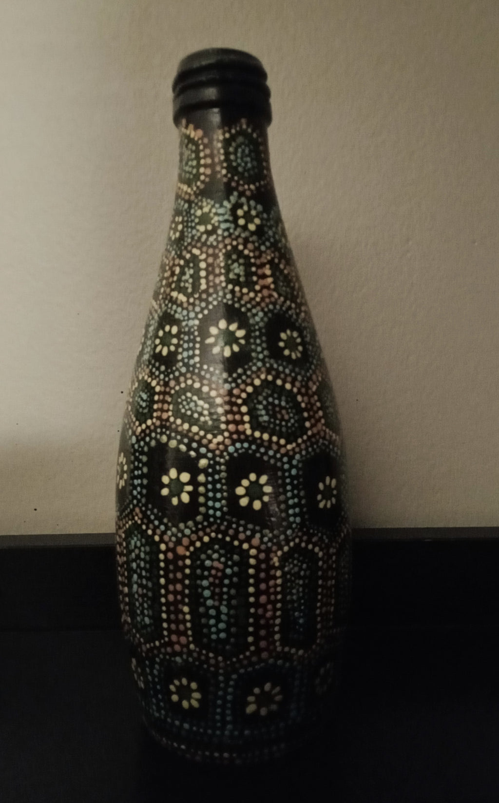 A black glass bottle decorated with intricate hand-painted Aboriginal-style dot art patterns in yellow, green, orange, and light blue.