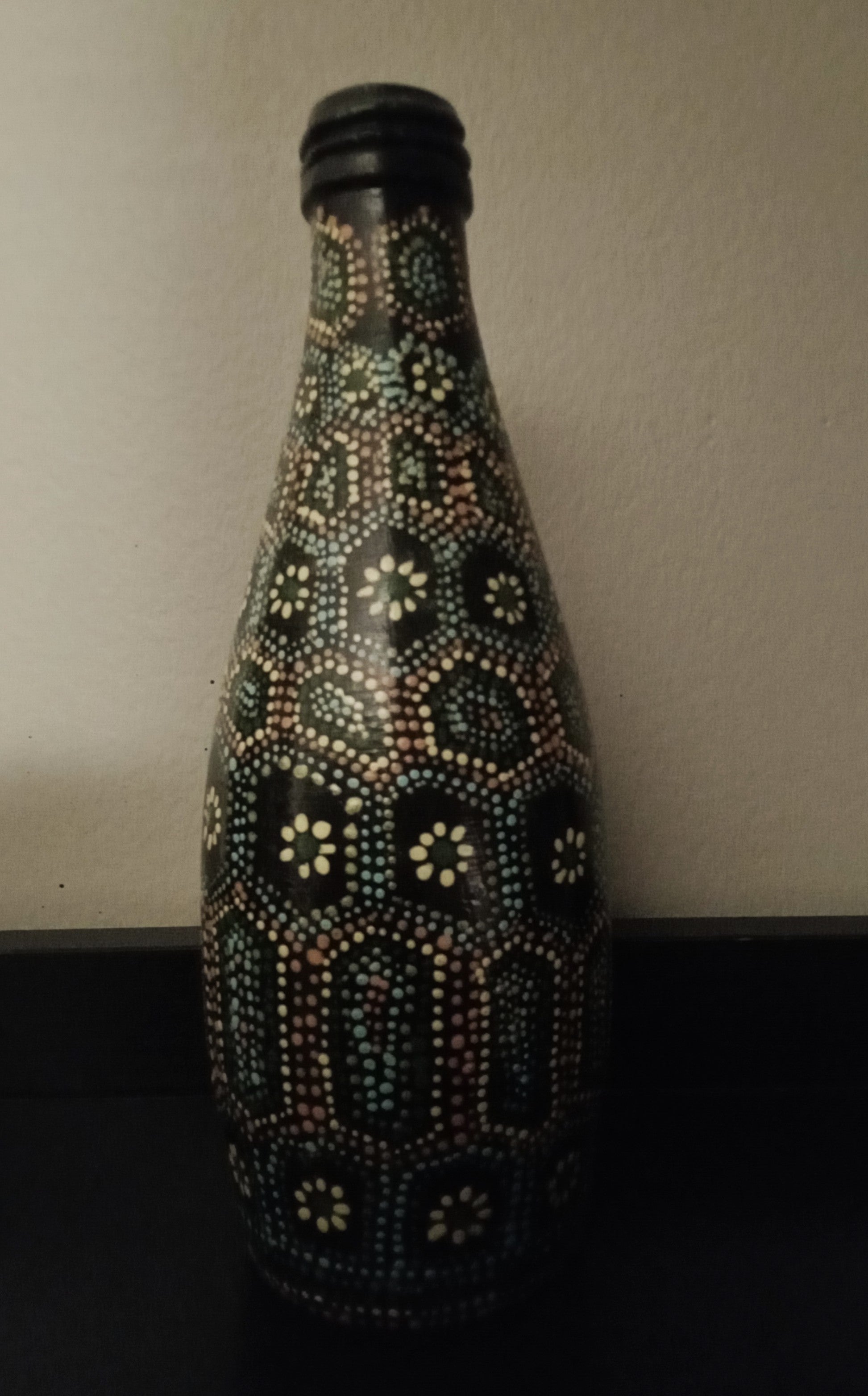 A black glass bottle decorated with intricate hand-painted Aboriginal-style dot art patterns in yellow, green, orange, and light blue.