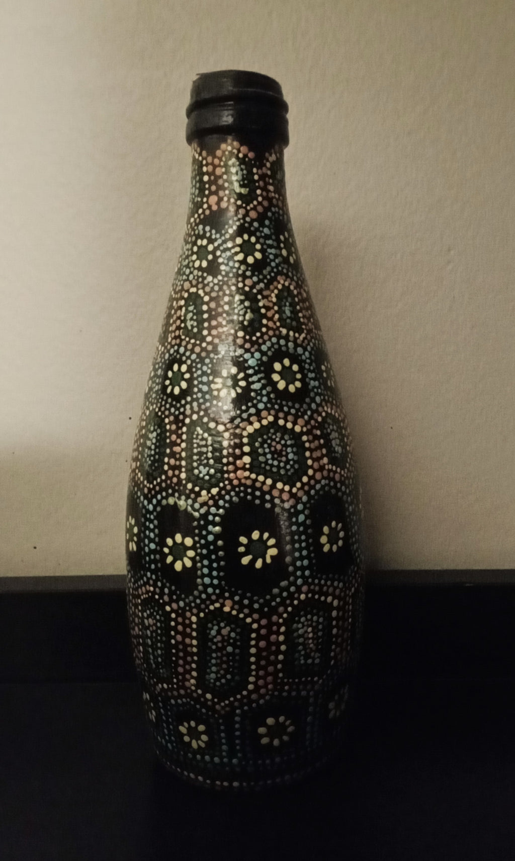 A black glass bottle decorated with intricate hand-painted Aboriginal-style dot art patterns in yellow, green, orange, and light blue.