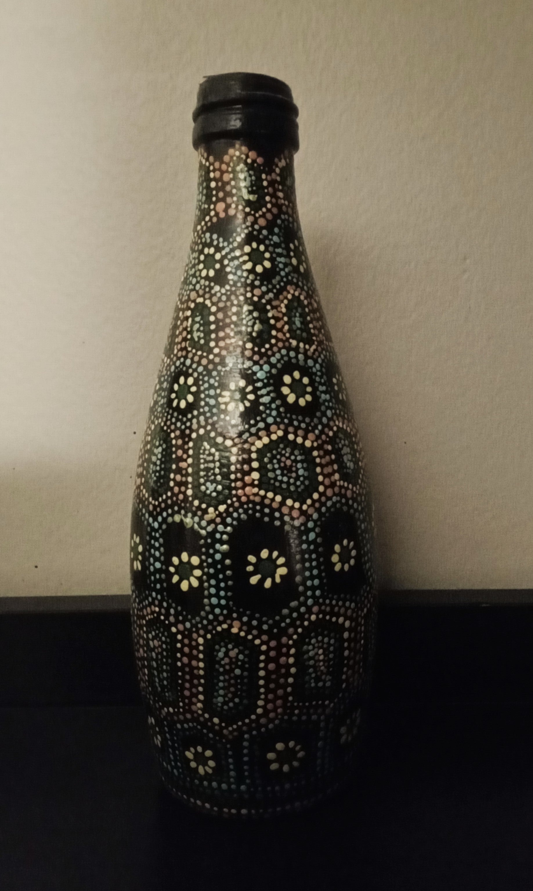 A black glass bottle decorated with intricate hand-painted Aboriginal-style dot art patterns in yellow, green, orange, and light blue.