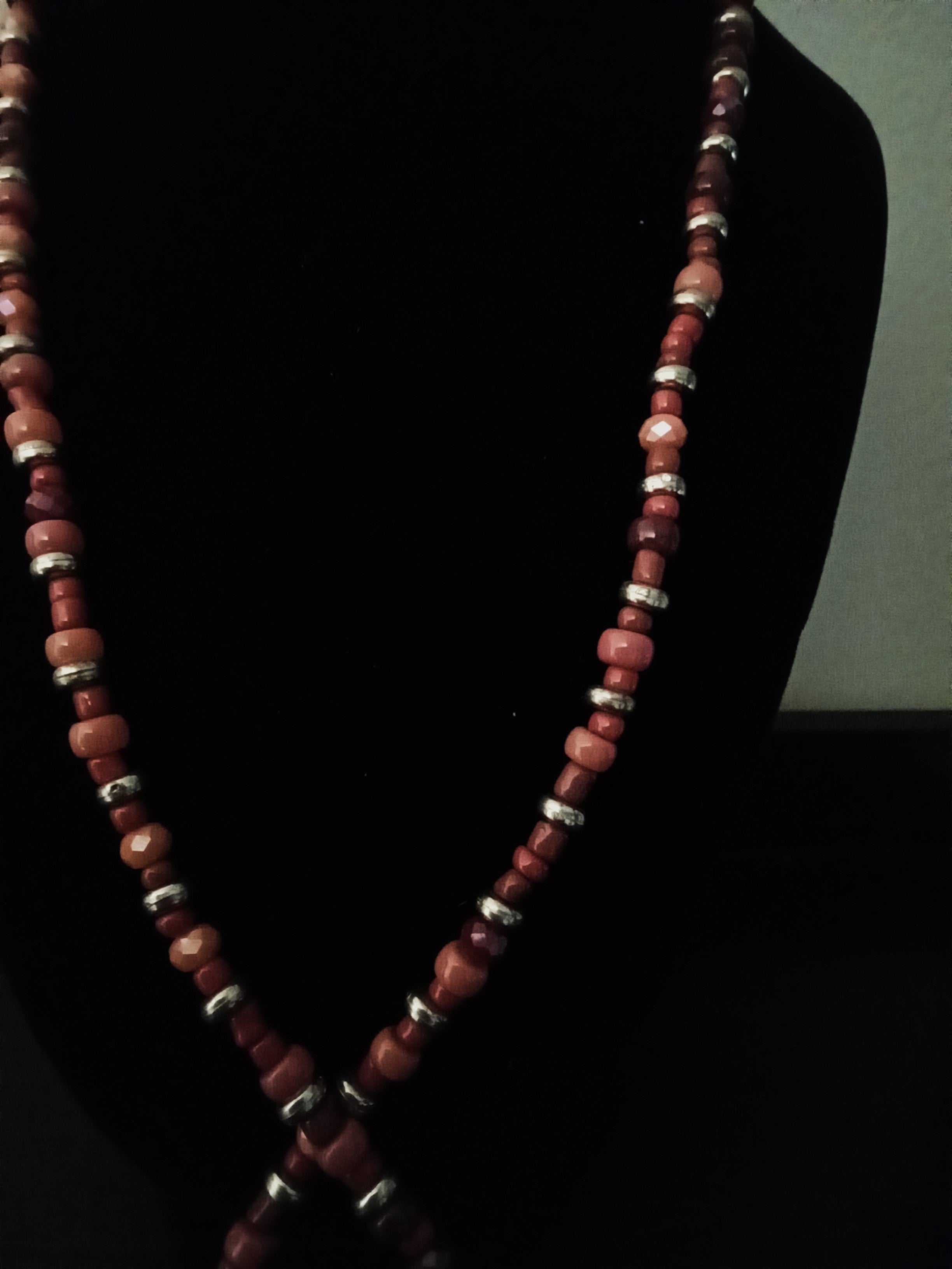 A beaded necklace made of reddish-brown stone beads and small golden spacer rings, displayed on a black jewelry bust against a dark background.