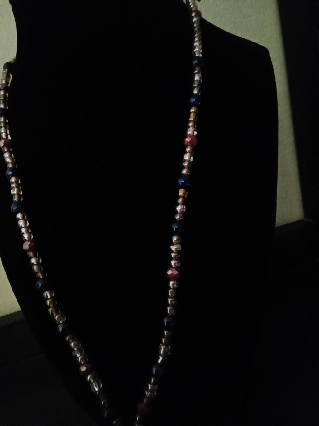 A long, beaded necklace displayed on a black jewelry bust against a dark background. The necklace features small iridescent spacer beads alternating with larger faceted beads in violet, pink, and maroon tones.