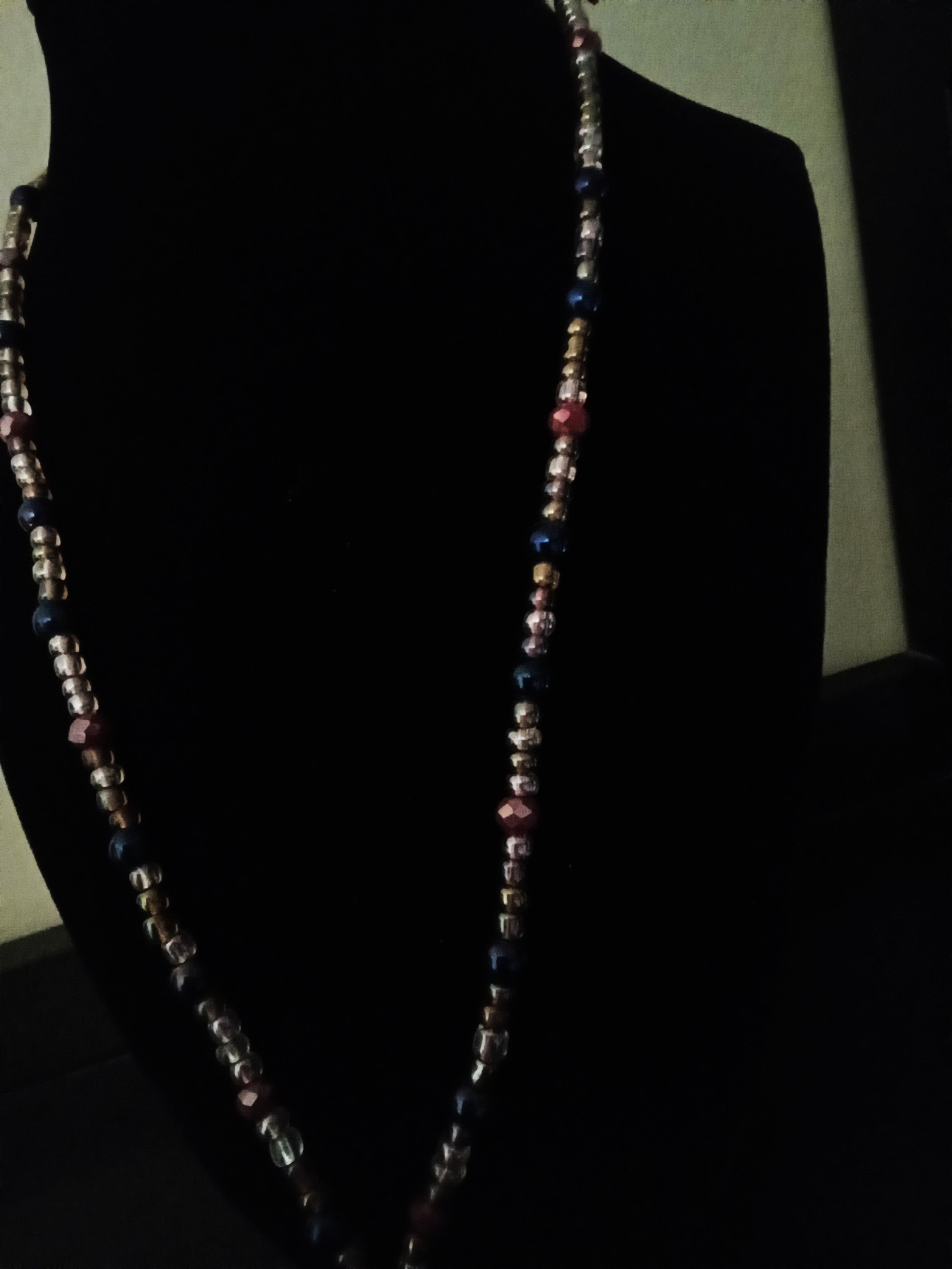 A long, beaded necklace displayed on a black jewelry bust against a dark background. The necklace features small iridescent spacer beads alternating with larger faceted beads in violet, pink, and maroon tones.